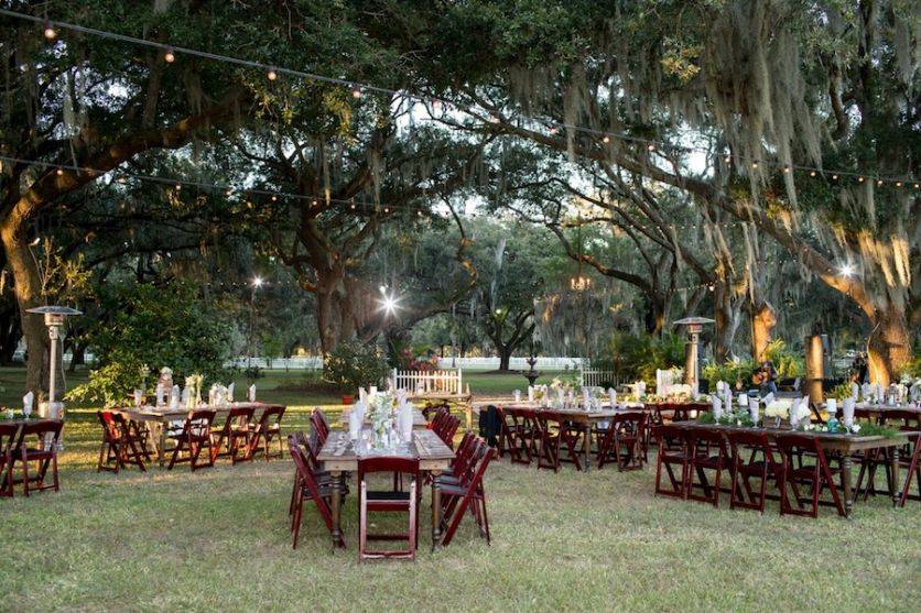 Outdoor farm event space with long tables, wooden chairs, and string lights set beneath large oak trees.